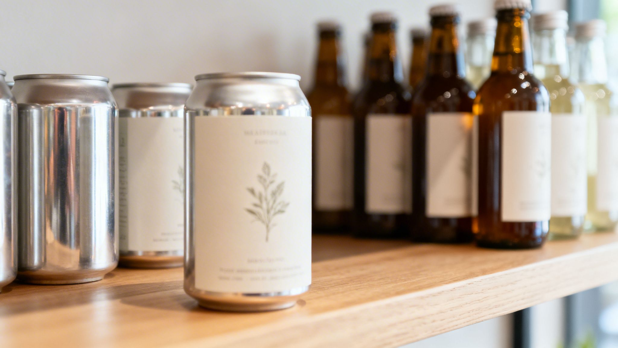 A close-up of aluminum cans and brown bottles with minimalist labels displayed on a wooden shelf.