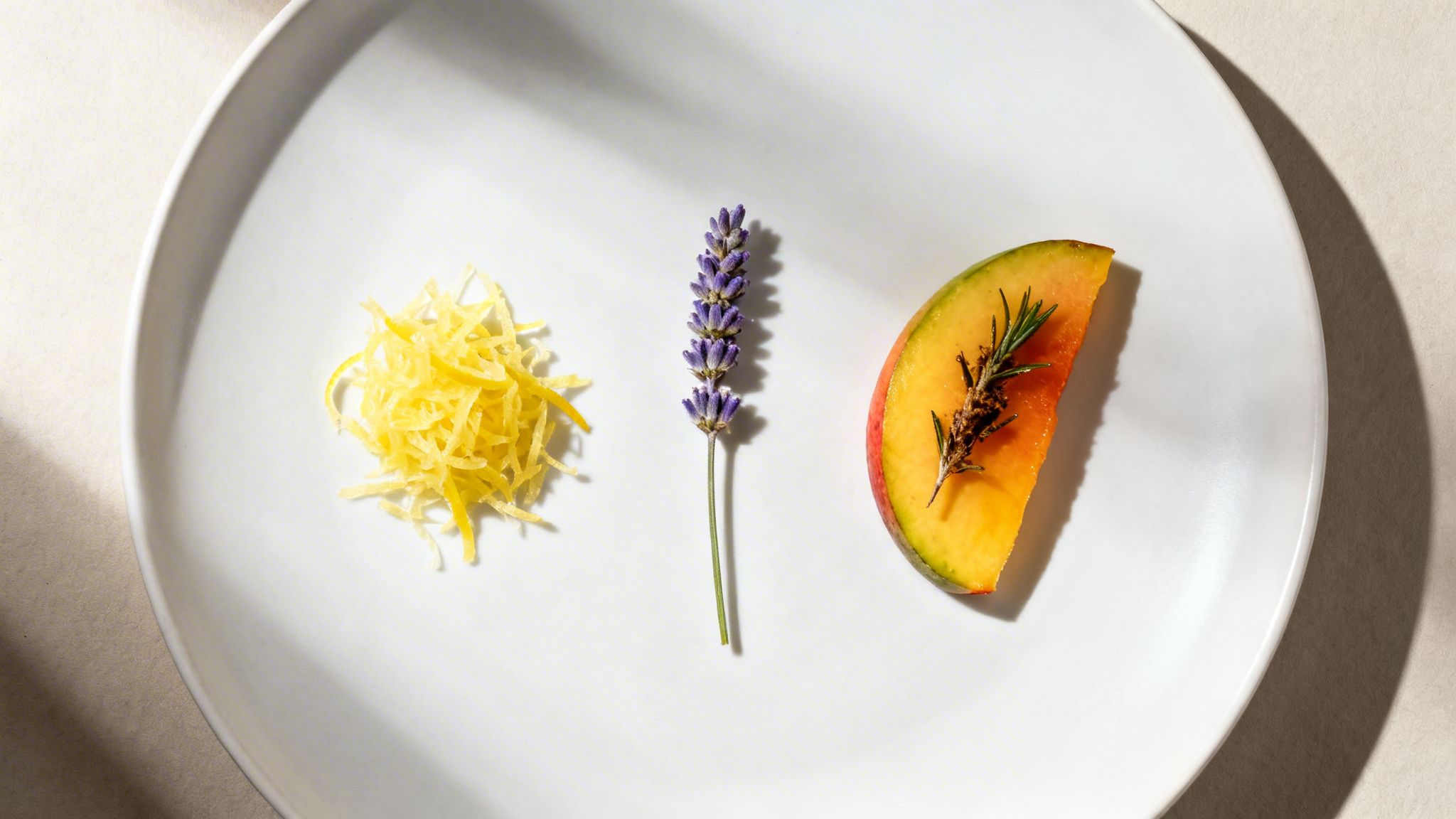 Lemon zest, lavender sprig, and mango slice with rosemary on a white plate.
