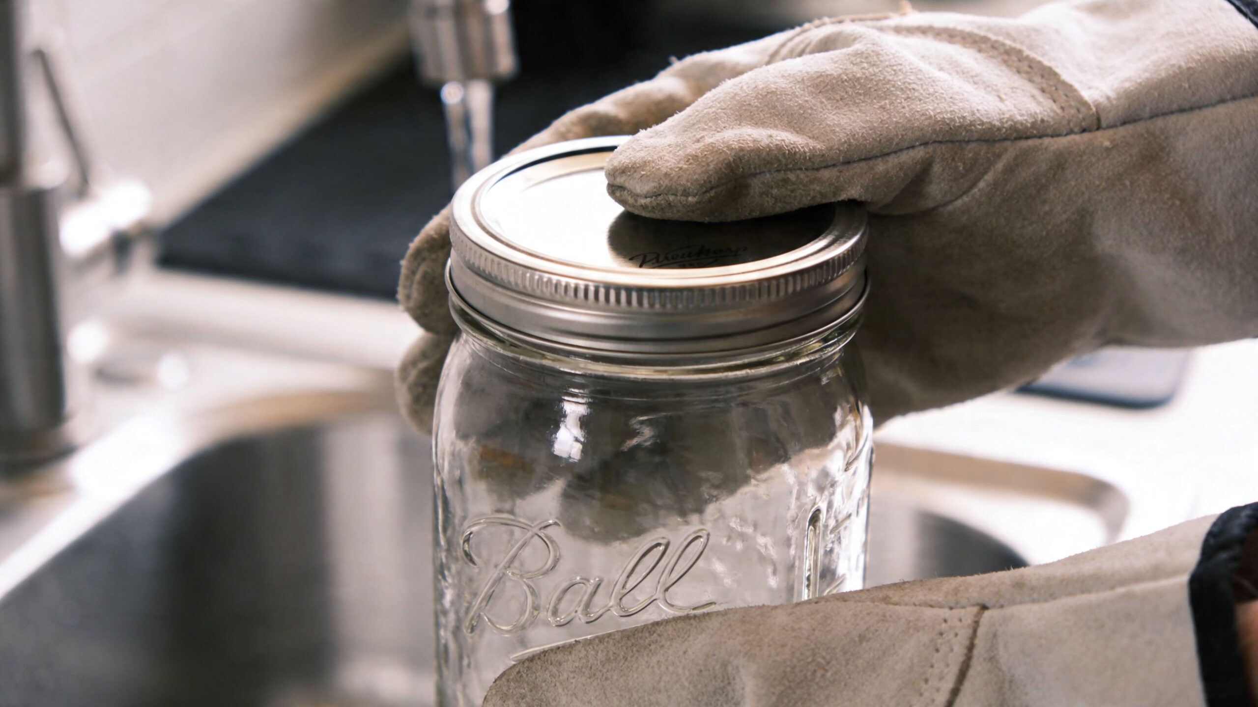 A person wearing heat-resistant leather gloves handling a glass mason jar in a kitchen sink setting.