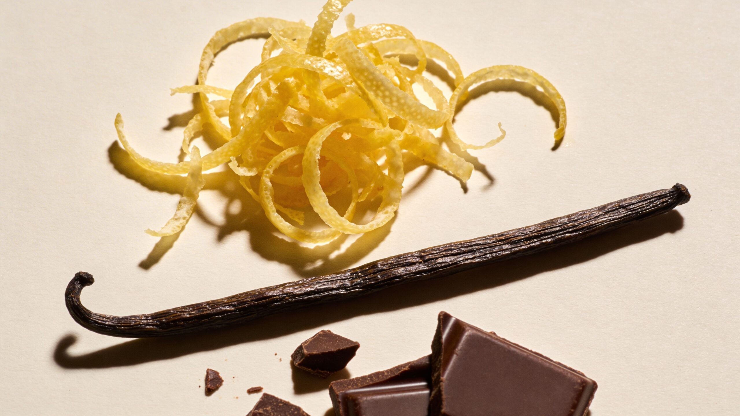 A fresh vanilla bean, lemon zest strips, and pieces of dark chocolate on a neutral background